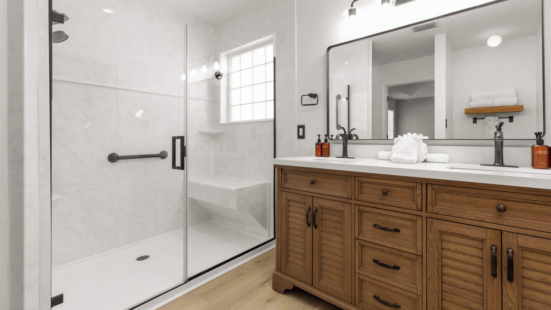 A modern bathroom with a glass shower enclosure, a wooden vanity with drawers, a large mirror, and a window providing natural light.
