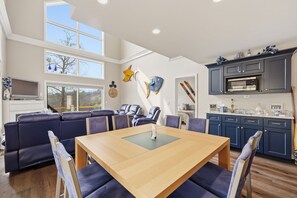 Ein geräumiges Wohnzimmer mit großem Fenster, ein Essbereich mit Holztisch und eine Küche mit dunklen Schränken und einer Mikrowelle.