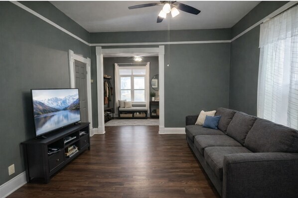 Un salon avec un téléviseur, un canapé, un ventilateur de plafond et une fenêtre avec des rideaux.