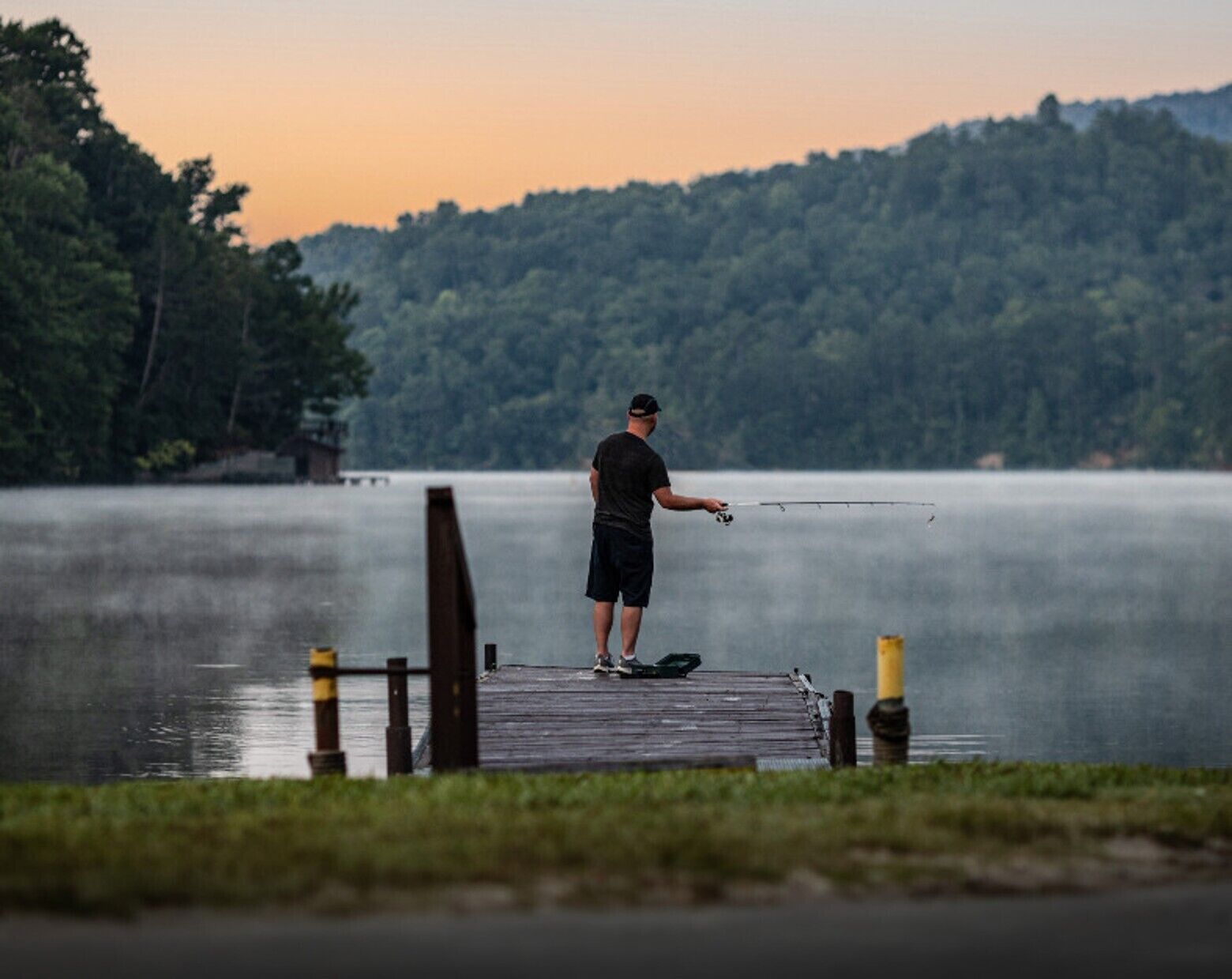 A person fishing on a wooden dock extending into a calm lake, surrounded by forested hills.