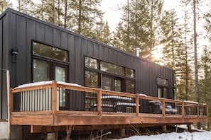 Una cabaña moderna con una terraza, ventanas amplias y un bosque de fondo.