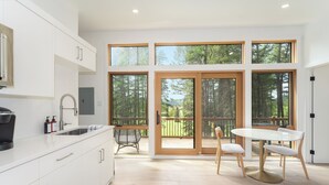 A modern kitchen with white cabinets, a coffee machine, and a dining area with a table and chairs. - North Star Ranch Modern Cabin by Beaver Lakes (Whitefish)