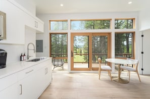 A modern kitchen with white cabinets, a coffee machine, and a dining area with a table and chairs. - North Star Ranch Modern Cabin by Beaver Lakes (Whitefish)