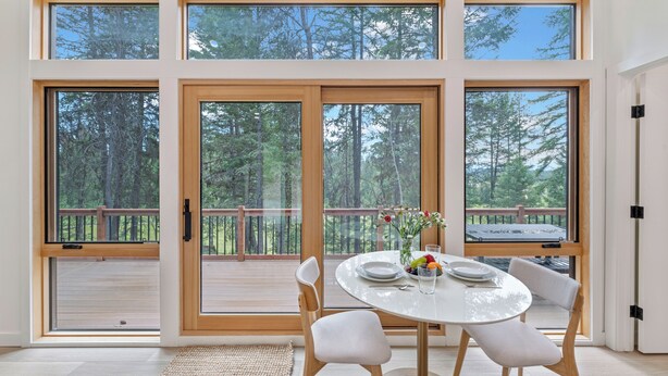 A dining area with a table, chairs, and a view of a forest through large windows. - North Star Ranch Modern Cabin by Beaver Lakes (Whitefish)