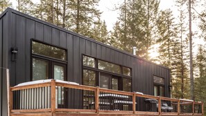 A modern cabin with a deck, large windows, and a forest backdrop. - North Star Ranch Modern Cabin by Beaver Lakes (Whitefish)
