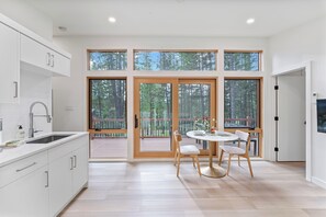 A modern kitchen with a large window, a dining area with a table and chairs, and a view of a deck. - North Star Ranch Modern Cabin by Beaver Lakes (Whitefish)