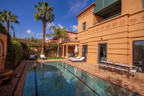 A swimming pool with lounge chairs and a dining area, surrounded by palm trees and a building with decorative patterns. - Golf villa amelkis 4 bedrooms (Marrakech)