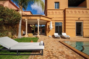 A poolside area with lounge chairs, a covered seating area, and a building with decorative patterns. - Golf villa amelkis 4 bedrooms (Marrakech)