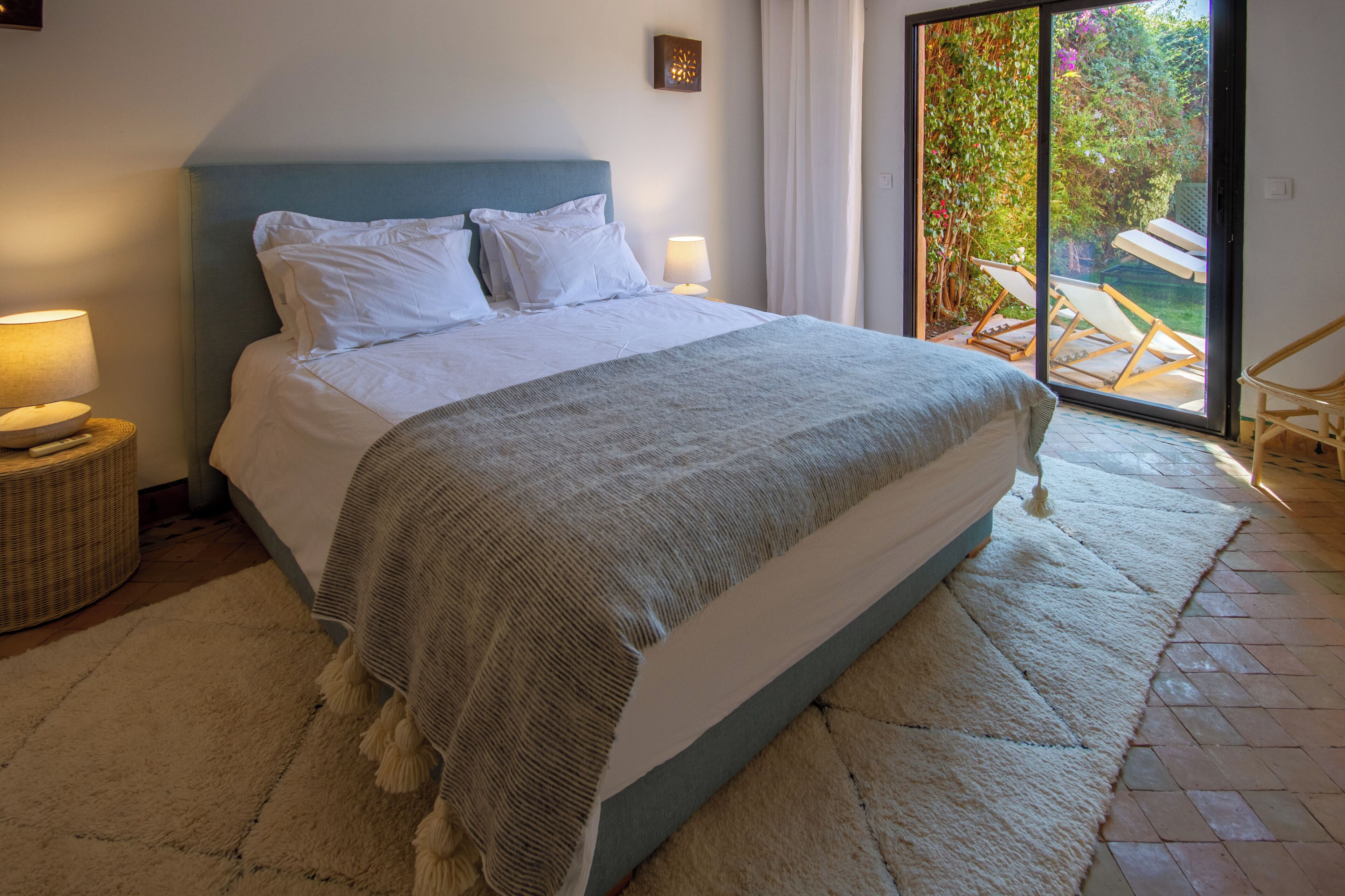 Un dormitorio con cama, lámparas de noche, una mesita de mimbre y vistas a un espacio exterior con una silla a través de un ventanal corredero.
