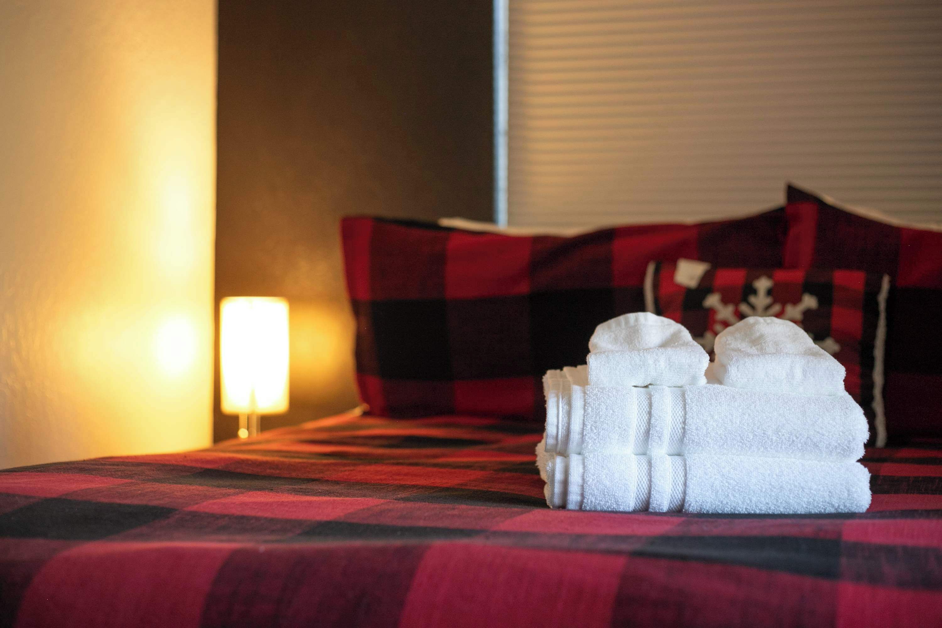 A bed with a red and black checkered bedspread, a white lamp, and a stack of white towels.
