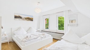 A bedroom with a white bed, a bedside table, a lamp, a radiator, and a window with a view of greenery.