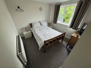 Un dormitorio con cama, mesita de noche, lámpara, ventana con cortinas, radiador y un mueble bajo para televisor con control remoto.