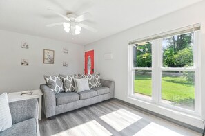 A modern living room with a grey sofa, patterned cushions, a white ceiling fan, and a large window with a view of a green lawn. - Bayou Villa w/ Dock, Kayaks & Fire Pit Fun (Pensacola)
