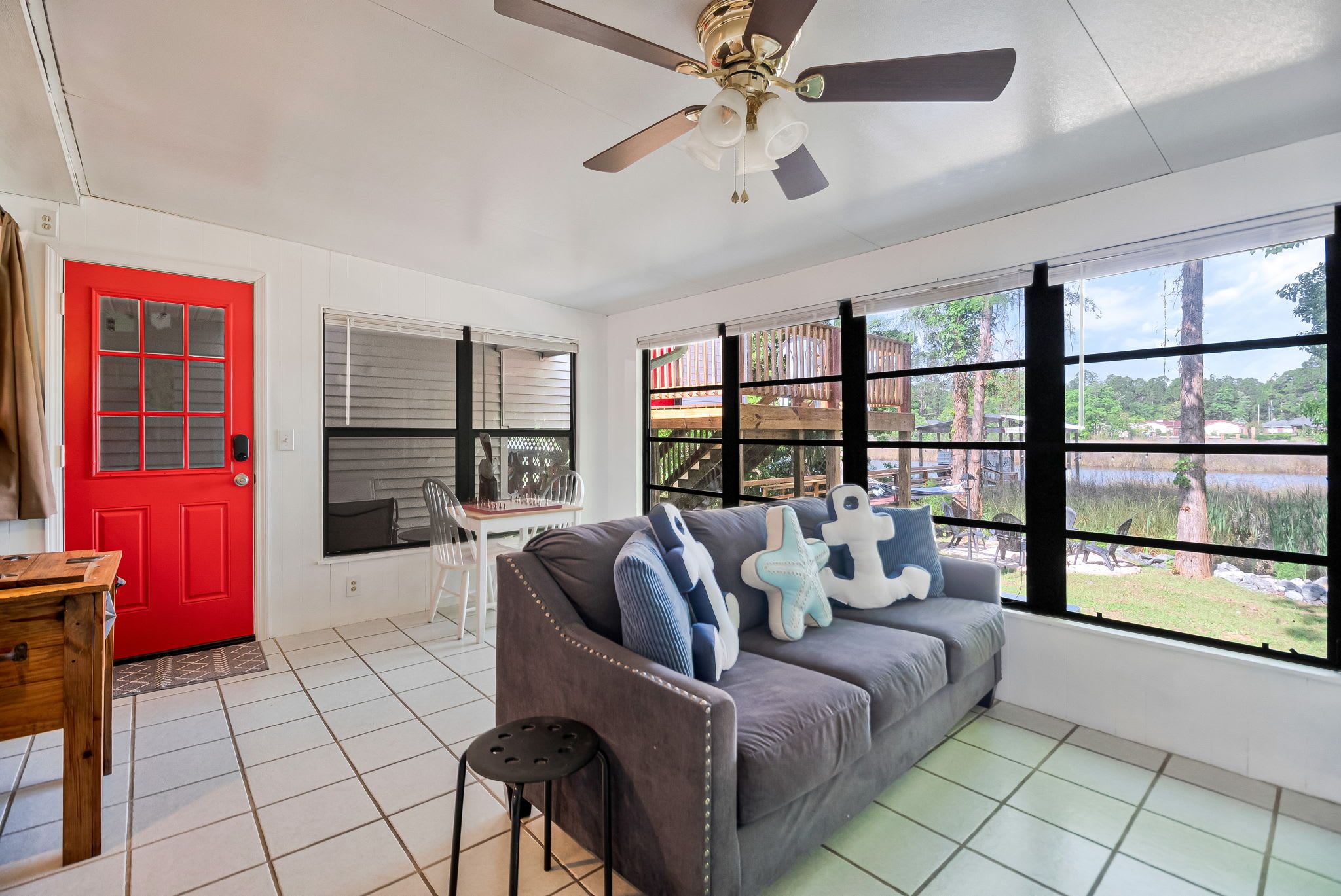 A room with a red door, a ceiling fan, a grey sofa with decorative pillows, a wooden table, and large windows with a view of a wooden deck and trees.