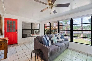 A room with a red door, a ceiling fan, a grey sofa with decorative pillows, a wooden table, and large windows with a view of a wooden deck and trees. - Bayou Villa w/ Dock, Kayaks & Fire Pit Fun (Pensacola)