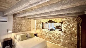 Een slaapkamer met een stenen muur, een bed met kussens, een nachtkastje, een spiegel en een houten plafond.