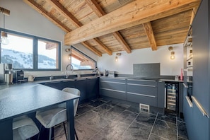 A modern kitchen with a stone tile floor, wooden ceiling beams, and a large window overlooking a snowy landscape. - Charming mountain chalet with jacuzzi & breathtaking view of the Aravis mountains (Le Grand-Bornand)