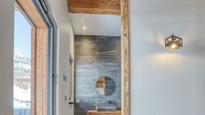 Une chambre avec une grande fenêtre, des poutres en bois au plafond, un mur en pierre, un banc en bois et un lit recouvert d’une couvre-lit à motifs.