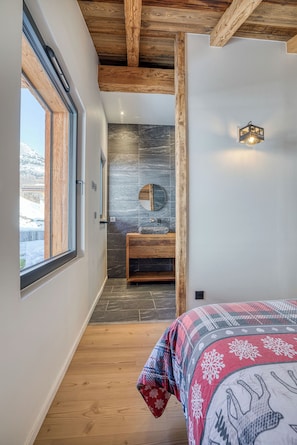 A bedroom with a large window, wooden ceiling beams, a stone wall, a wooden bench, and a bed with a patterned bedspread. - Charming mountain chalet with jacuzzi & breathtaking view of the Aravis mountains (Le Grand-Bornand)