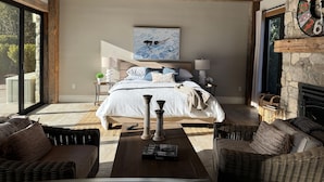 A bedroom with a bed, two bedside tables, a ceiling fan, a fireplace, and a large window.