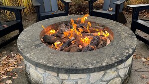 A stone fire pit with logs, surrounded by four black Adirondack chairs.
