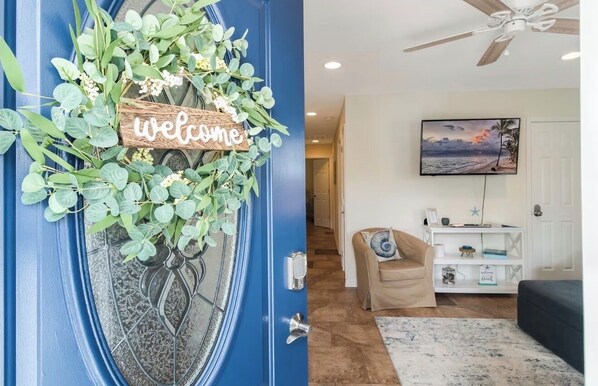 A blue door with a welcome sign, a television, a beige sofa, and a ceiling fan.