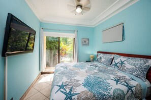 Ein Schlafzimmer mit Bett, Deckenventilator, Fernseher und einem Fenster mit Blick auf Grünflächen.