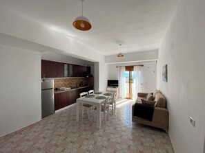 Eine moderne Küche mit dunklen Schränken, ein Essbereich mit weißem Tisch und Stühlen sowie eine Wohnbereits mit einer Couch und einem Fernseher.