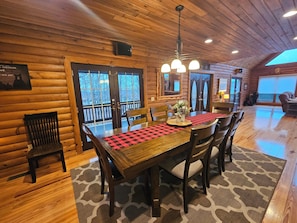 A wooden interior with a dining table, chairs, a chandelier, and a large window.