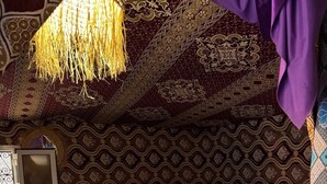 Una habitación con alfombra estampada, cama con manta morada, lámpara colgante y pared con diseños intrincados.