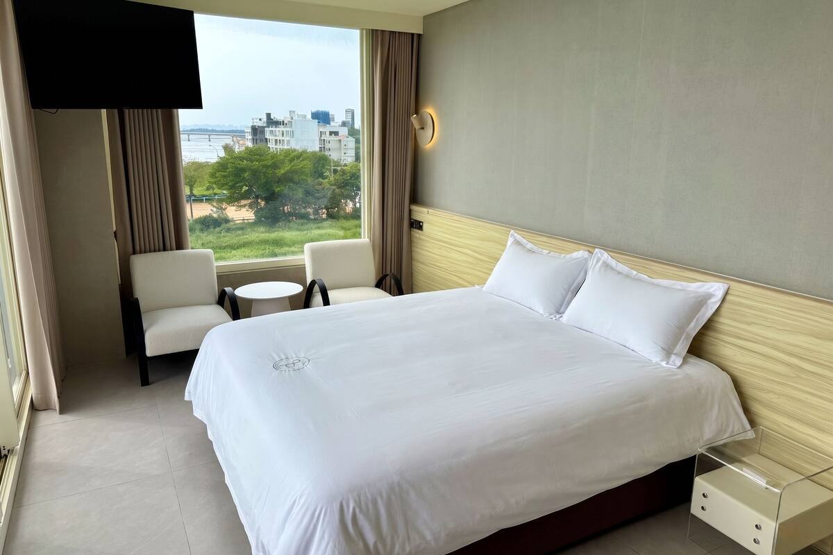 Habitación de hotel con una cama grande, un televisor montado en la pared, una mesita, dos sillas y vistas a la ciudad desde la ventana.