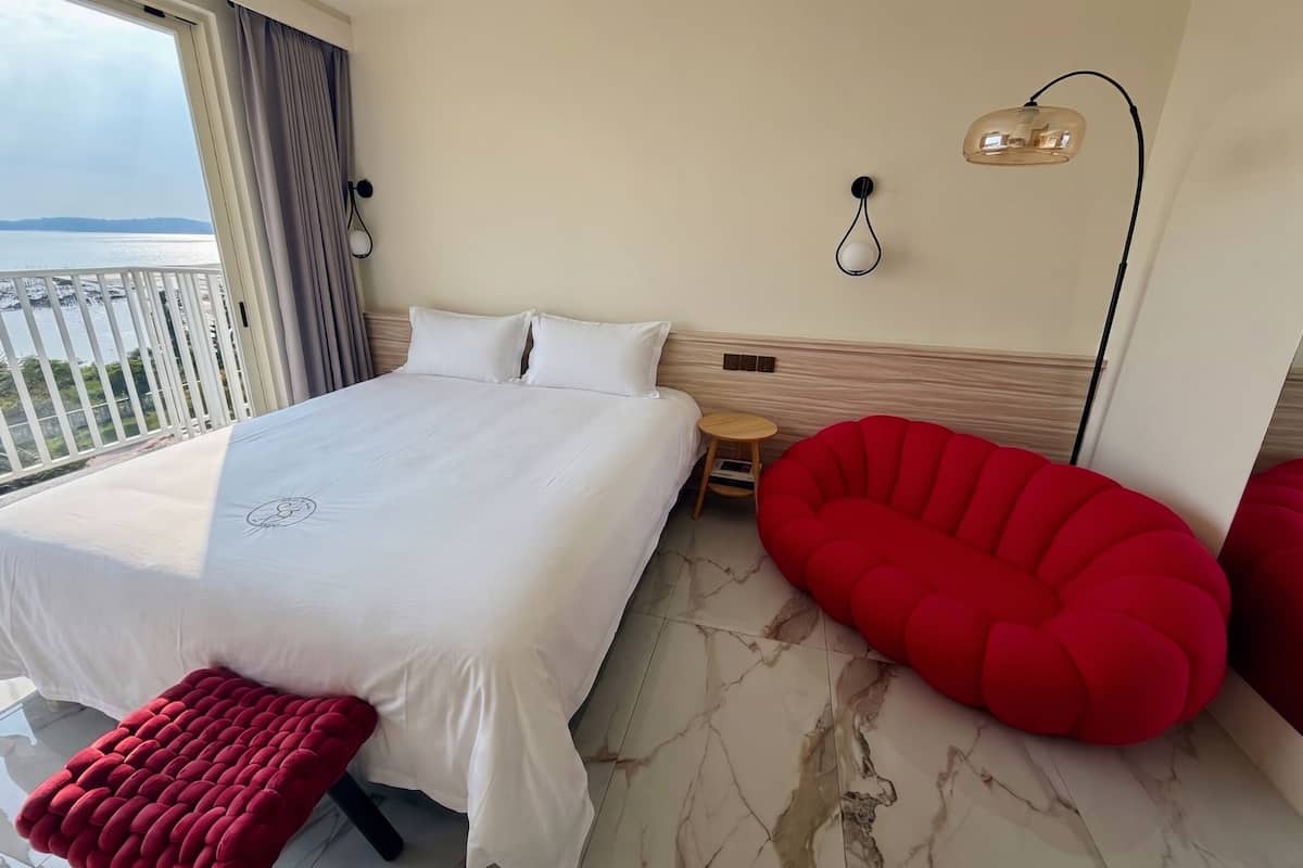 Habitación de hotel con una cama, una mesita redonda pequeña, una silla roja, una lámpara de pared y vistas al mar.