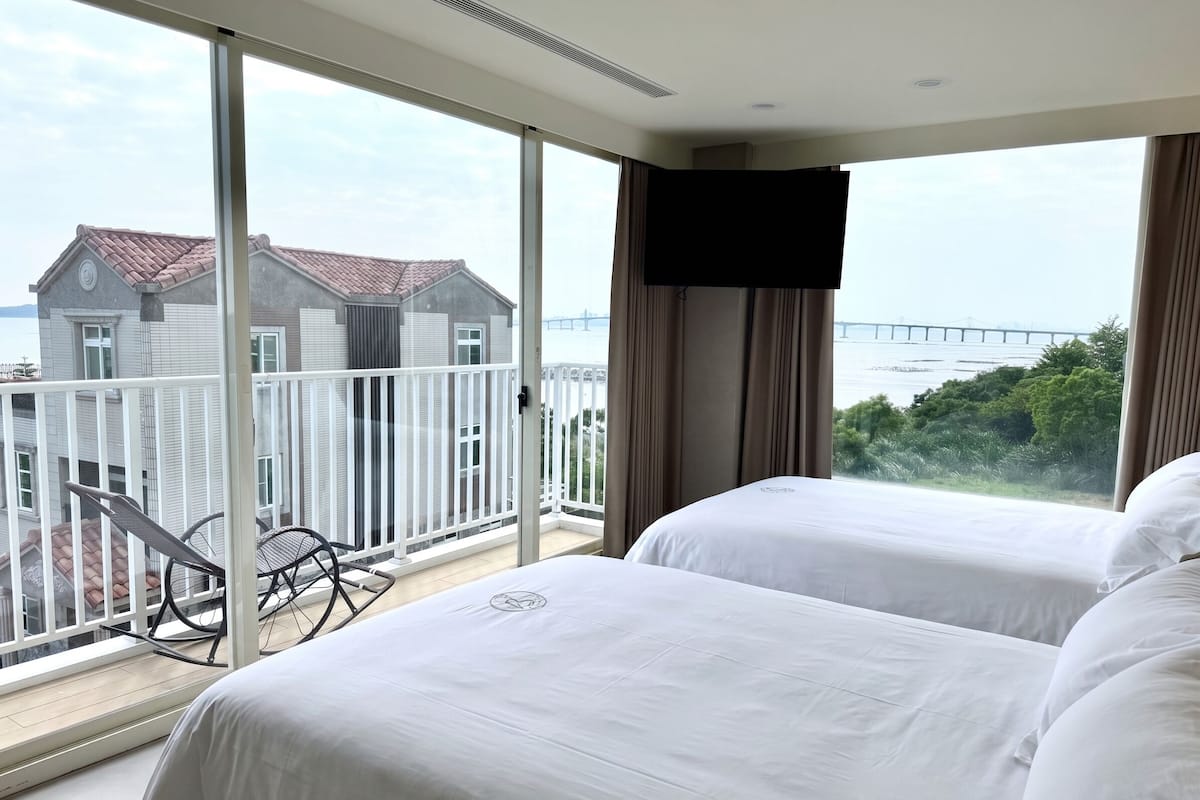 Habitación de hotel con dos camas, un balcón con una silla y vistas a un puente y edificios.