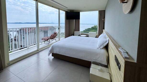 A hotel room with a large bed, a TV, a desk with a lamp, a chair, and a view of the sea. - CP Tide whisper (Jinning)