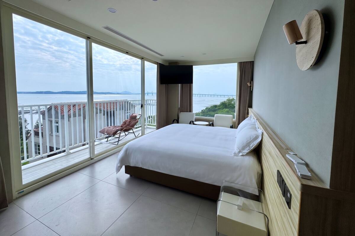 Habitación de hotel con una cama grande, televisor, escritorio con lámpara, silla y vista al mar.