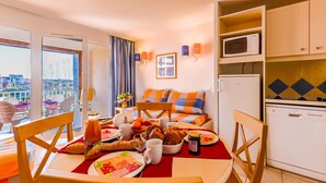 A room with a dining table set with food, a sofa with colorful cushions, a kitchen area with appliances, and a view through a window. - Mediterranean Escape in Le Barcares (Le Barcares)