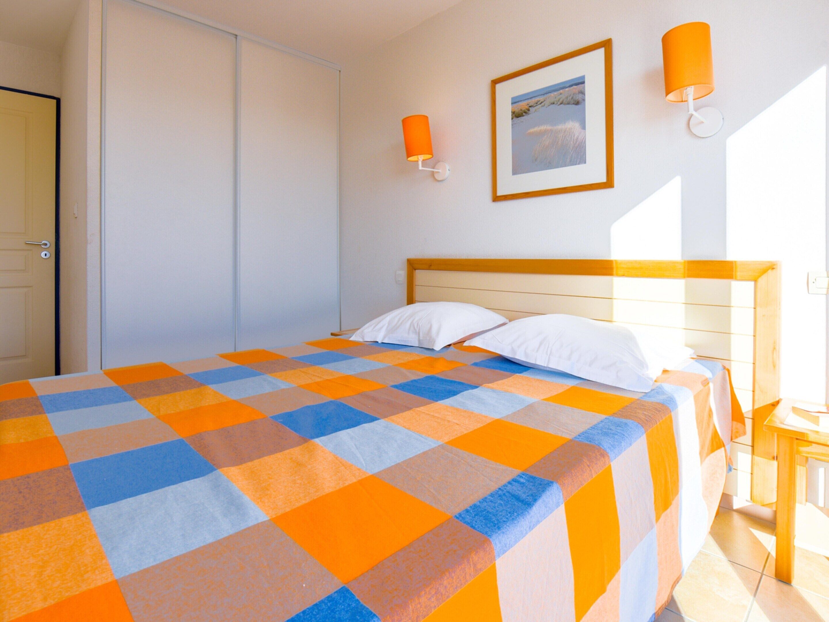 A bedroom with a checkered bedspread, a wooden headboard, two white pillows, a framed picture on the wall, and two wall-mounted lamps.