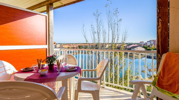 A balcony with a table set for dining, chairs, and a view of the sea and buildings. - Mediterranean Escape in Le Barcares (Le Barcares)