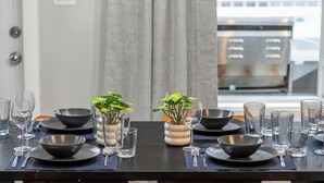 A dining table set with multiple bowls, glasses, and cutlery, with potted plants and a view of a building through a window.