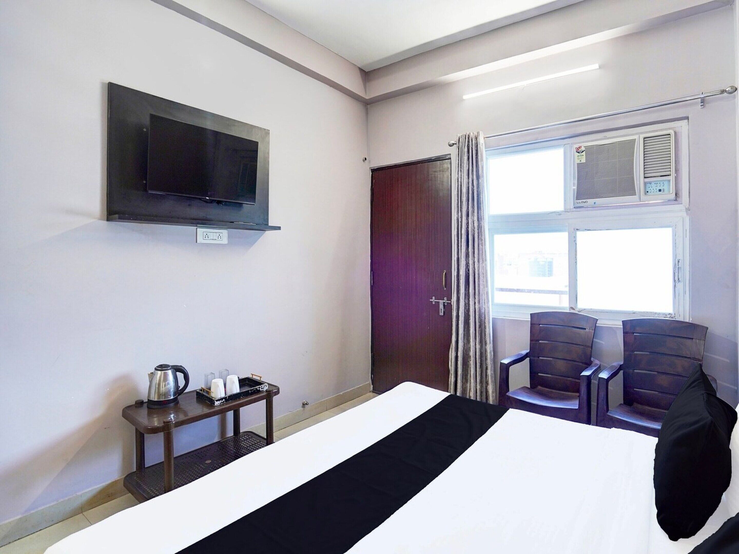 A hotel room with a bed, a TV mounted on the wall, a small table with a kettle and cups, two chairs, and a window with curtains.