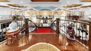 A grand interior with a central staircase, ornate ceiling, and a red carpet leading to a second-floor balcony.