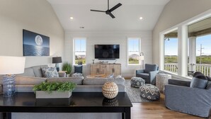 A spacious living room with a large flat-screen TV, a ceiling fan, and a view of the outdoors through large windows. - Sunset State of Mind: Private Pool | Hot Tub | Elevator (Nags Head)