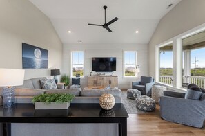 A spacious living room with a large flat-screen TV, a ceiling fan, and a view of the outdoors through large windows. - Sunset State of Mind: Private Pool | Hot Tub | Elevator (Nags Head)