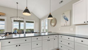 A kitchen with white cabinets, a large island, pendant lights, and artwork on the walls. - Sunset State of Mind: Private Pool | Hot Tub | Elevator (Nags Head)