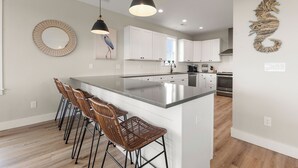 A modern kitchen with white cabinets, a large island with bar stools, pendant lighting, and a decorative wall piece. - Sunset State of Mind: Private Pool | Hot Tub | Elevator (Nags Head)