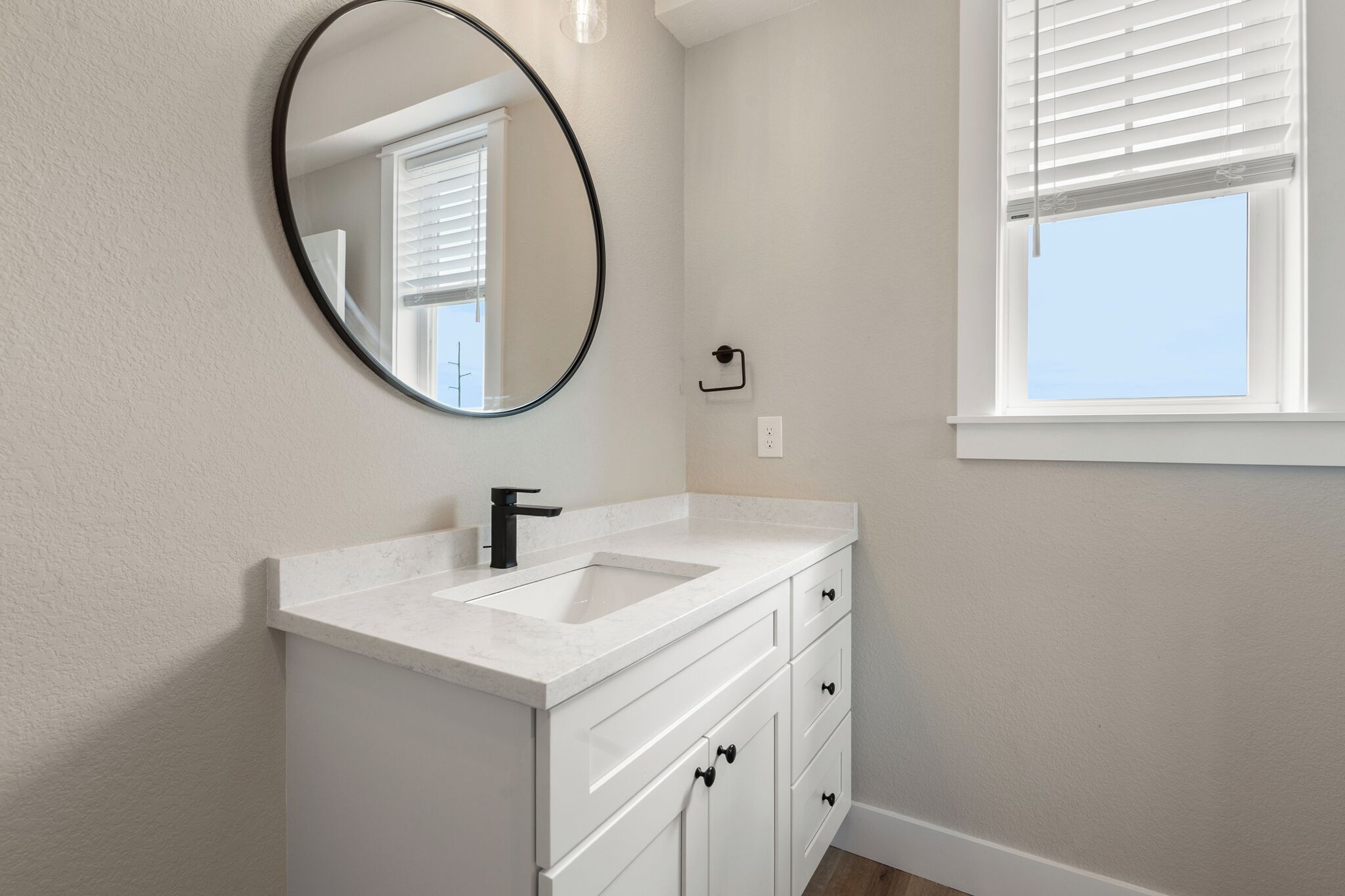 A bathroom with a white vanity, a round mirror, a sink, and a window with blinds.
