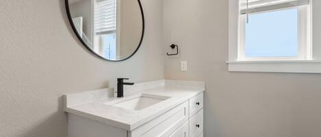 A bathroom with a white vanity, a round mirror, a sink, and a window with blinds.