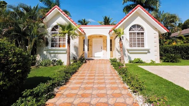 Una casa de dos pisos con una entrada central, rodeada de abundante vegetación y palmeras.
