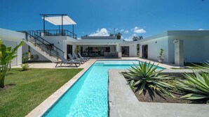 Un área moderna junto a la piscina con una piscina azul cristalina, un espacio de estar cubierto al aire libre y una escalera que lleva a un segundo nivel.