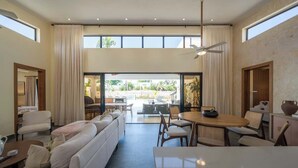 Amplia sala de estar con sofá, mesa de comedor y sillas, ventanas grandes y vistas al exterior.